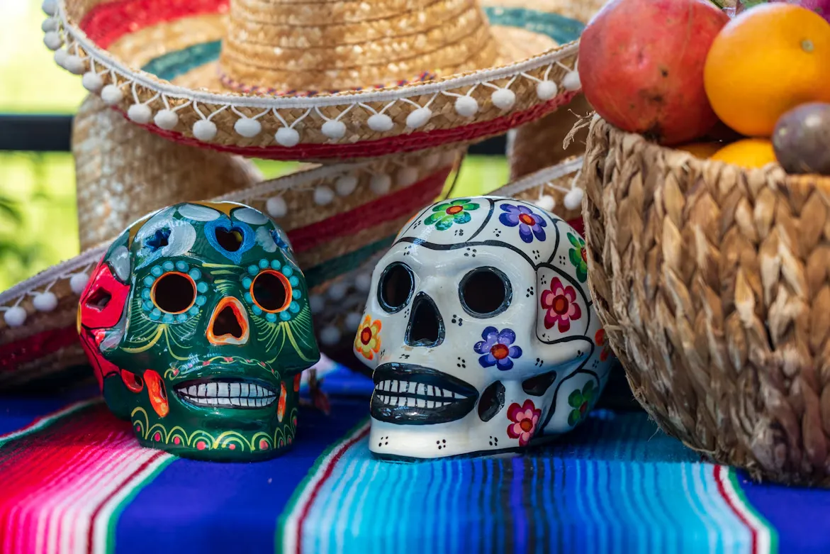 Two painted sugar skulls on a colorful striped blanket, with straw hats and a basket of fruit behind them