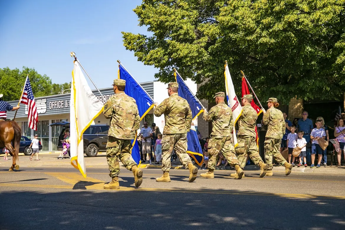 A color guard carrying flags marches down a sunny street while families watch from the curb at the Gaithersburg Labor Day Parade