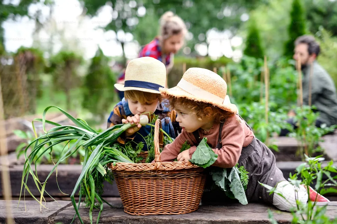 Two young children in straw hats leaning over a wicker basket of vegetables on a farm, with adults working behind them