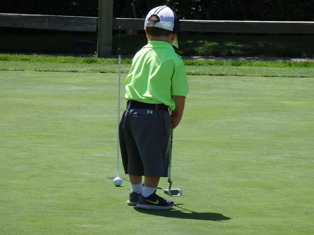 A little boy in a green shirt and cap holding a golf club on a putting green