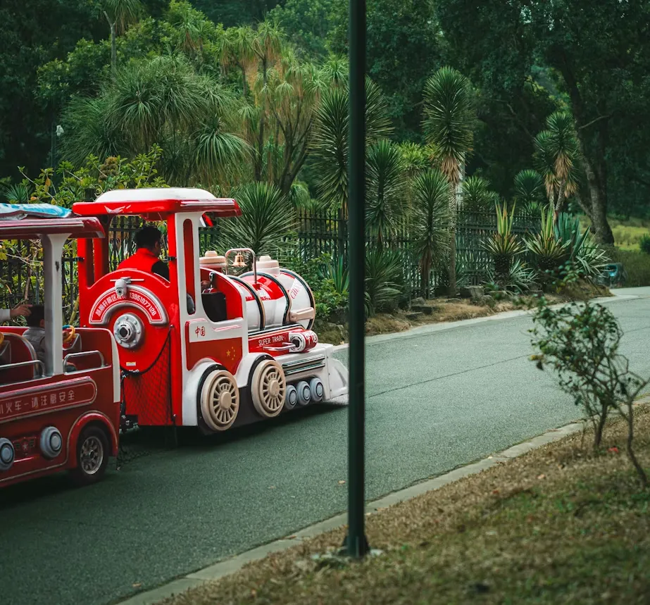 A small red and white miniature train carrying riders along a paved park path