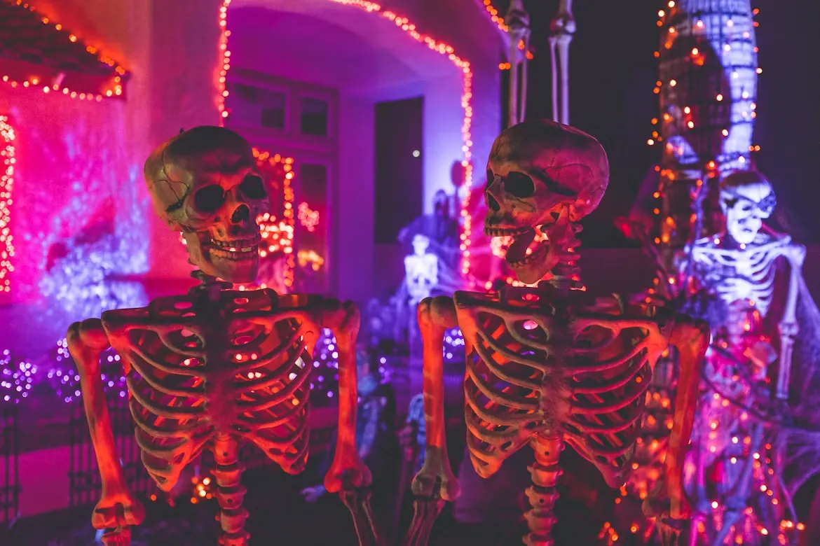 Two skeleton figures lit by pink and orange string lights on a decorated porch at Halloween