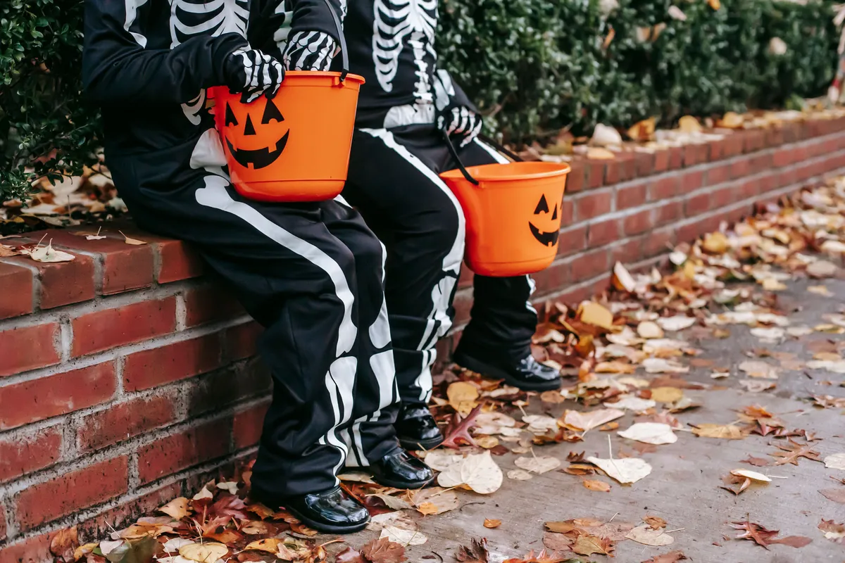 Two kids in skeleton costumes sitting on a brick wall with jack-o-lantern candy buckets