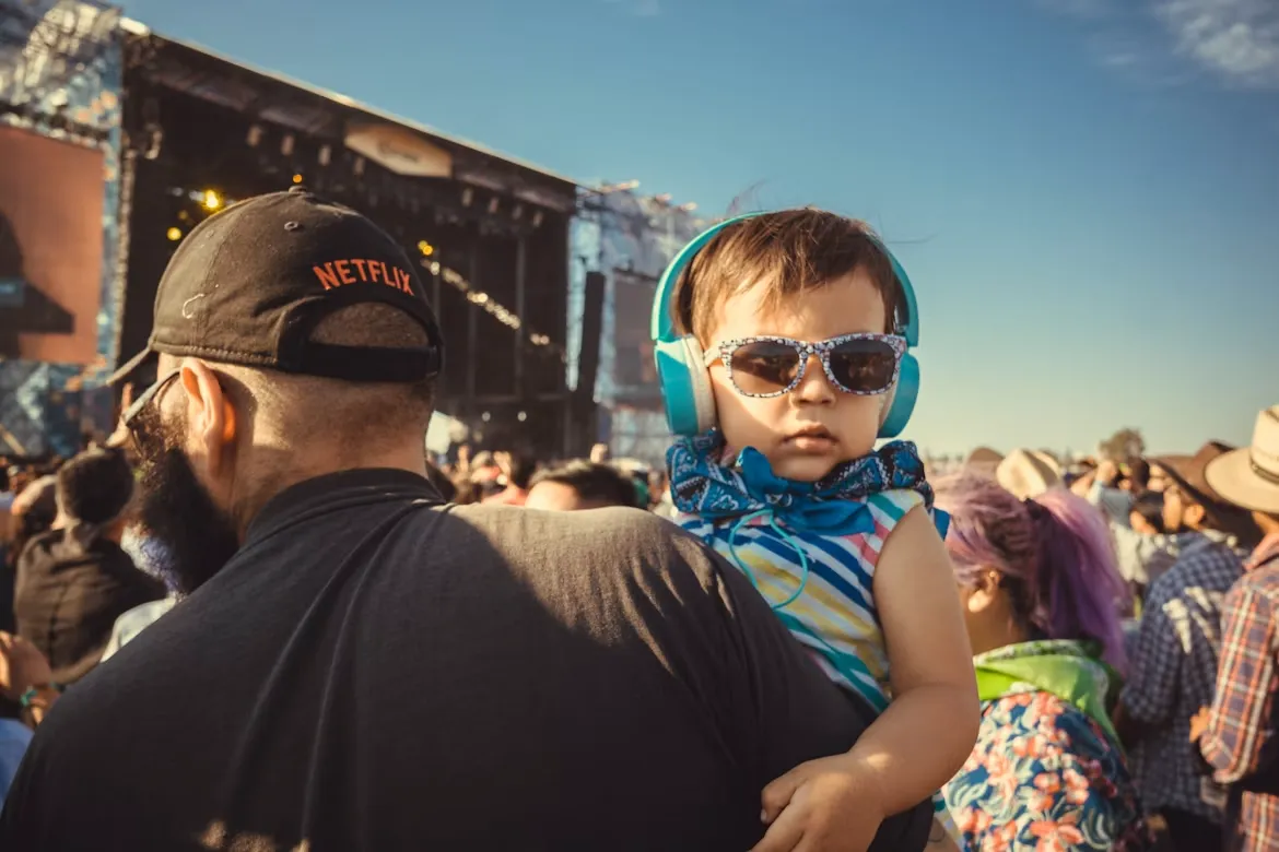 A toddler in sunglasses and ear protection headphones on a parent's shoulders at an outdoor concert