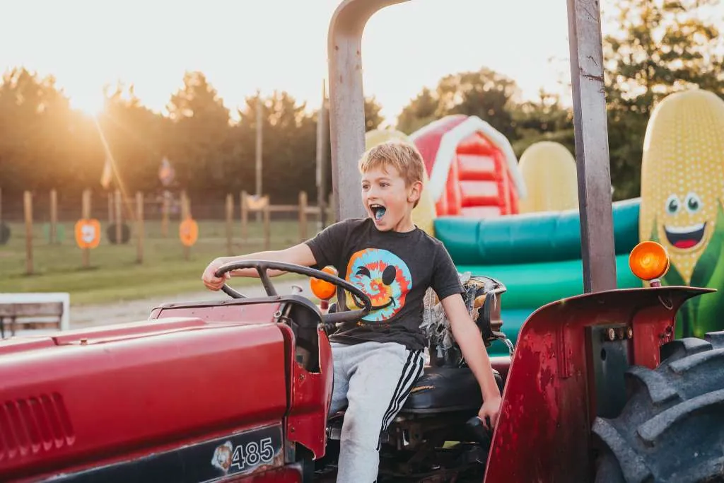 A boy grinning in the seat of a red tractor at a fall festival, with bounce houses and a giant corn balloon behind him