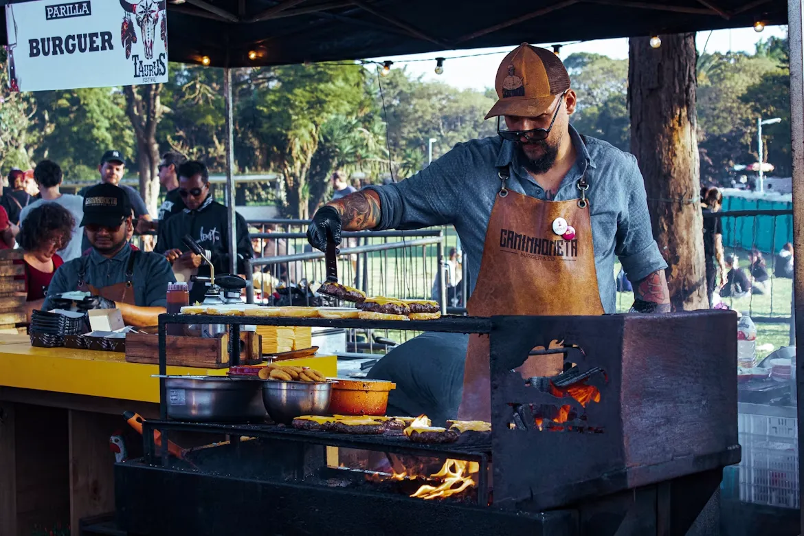 A cook grilling food at an outdoor food stand with people lined up behind him